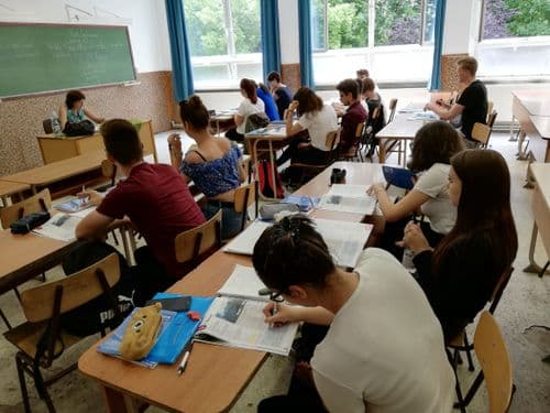 tantermi kép felnőtt tanulókkal, akik padokban ülnek, jegyzetelnek vagy hallgatják a tanárnőt, aki a tábla előtt ül a tanári asztalnál