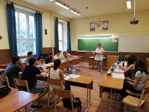 tantermi kép, tanulók körban a padoknál ülnek, a tanárnő előttük teleírt hagyományos krétás tábla előtt állva magyaráz, kezében nyitott könyv.