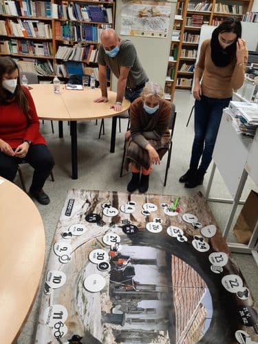 Társasjáték a szentendrei lépj feljebb projektünk keretében. Egy nagy asztalon van a játéktábla, mellette a játékosok elgondolkodva nézik és maszkot viselnek a covid miatt