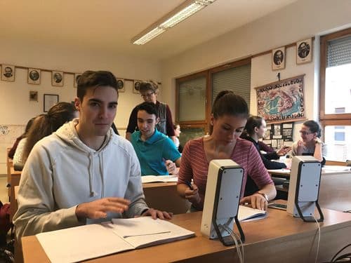 férfi, női tanulók vegyesen ülnek a padban, előttük számítógép-hangfalak, amit hallgatnak és jegyzetelnek