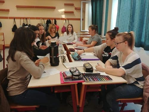  10 lánytanuló egy asztalnál ül, rajzol, ír valamit előttük tollak, színes filcek, stb.