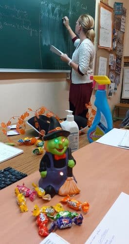 Tanárnő ír a táblára, háta mögött az íróasztalán vicces halloween boszokányfigura ül, körülötte cukorkák
