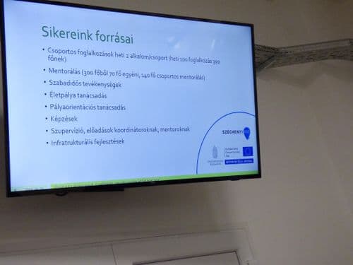 Nagyméretű fali TV a DKA aulában, rajta éppen a "Sikereink forrása.." című szöveg megy.
