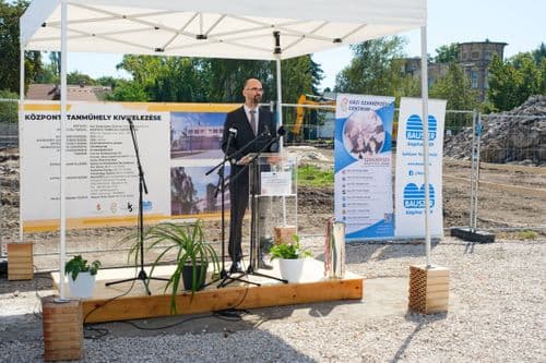 Az államtitkár az emelvényen beszél, mögötte az építési terület és a látványterv plakátok